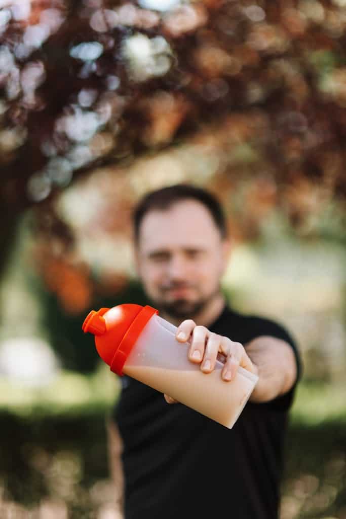 Man holding out a protein shake bottle outdoors, wearing casual black shirt.