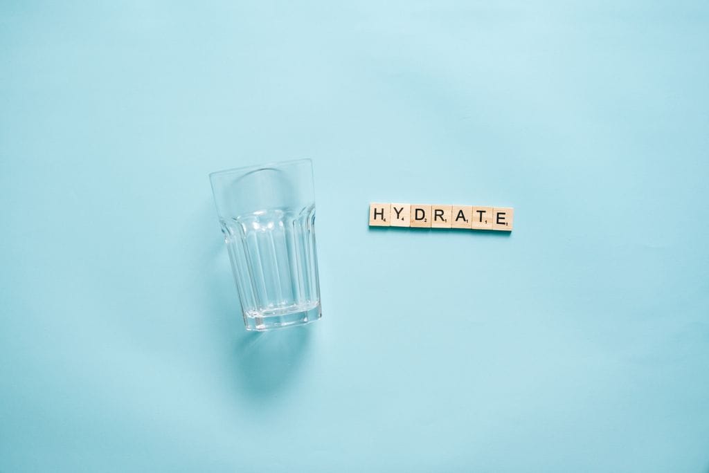 Minimalist flat lay of a drinking glass and letter tiles spelling 'HYDRATE' against a blue background.