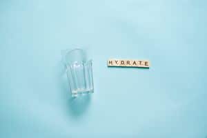Minimalist flat lay of a drinking glass and letter tiles spelling 'HYDRATE' against a blue background.
